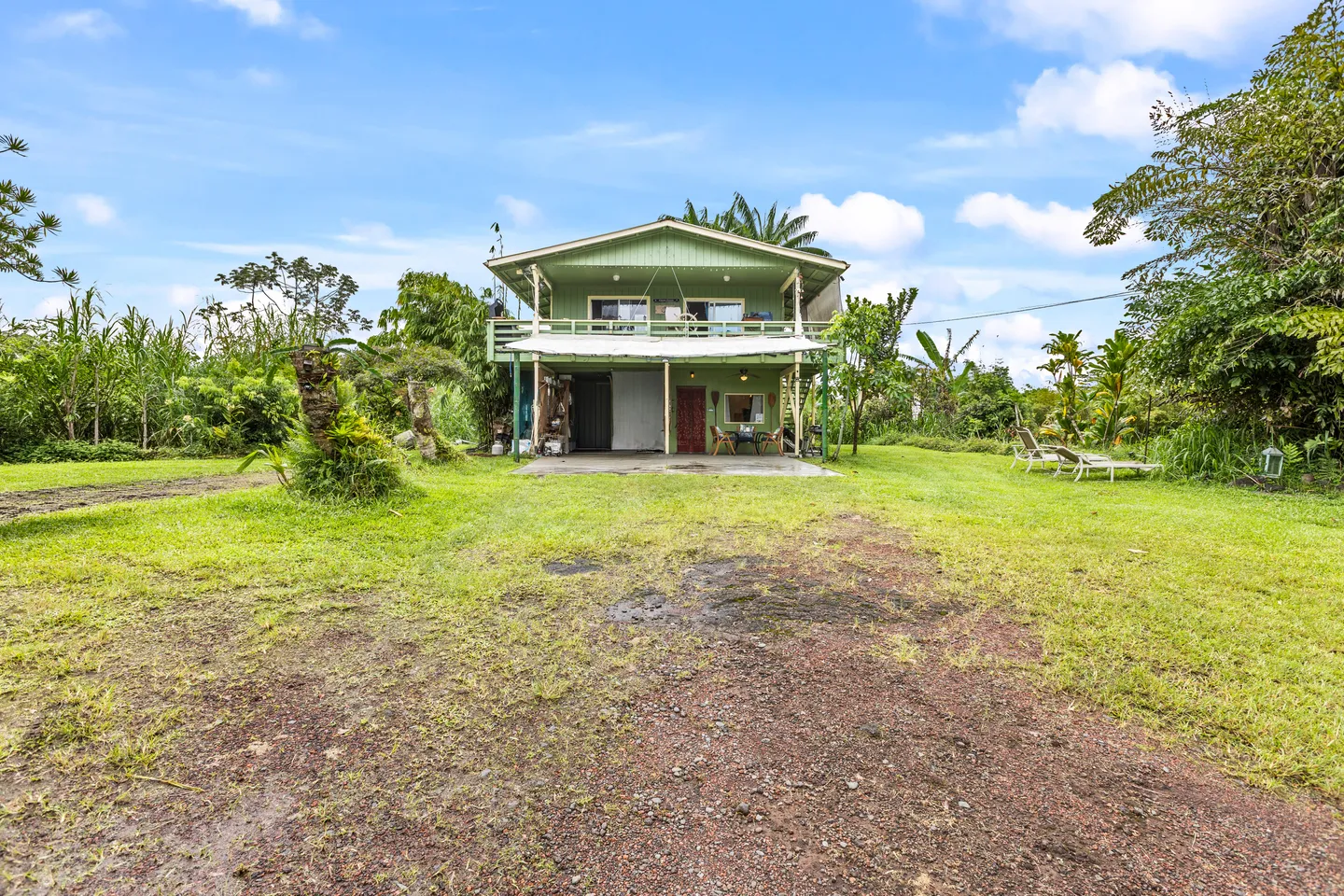 Exterior view of a two-story light green home surrounded by a grassy lawn and tropical foliage under a blue sky.