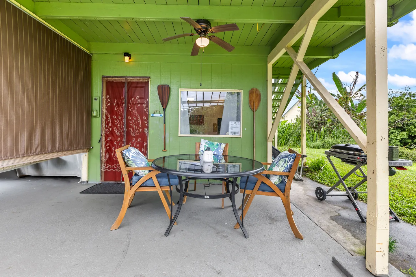 Covered outdoor patio with a round glass-top dining table, wooden chairs with blue cushions, a ceiling fan, and a grill nearby.