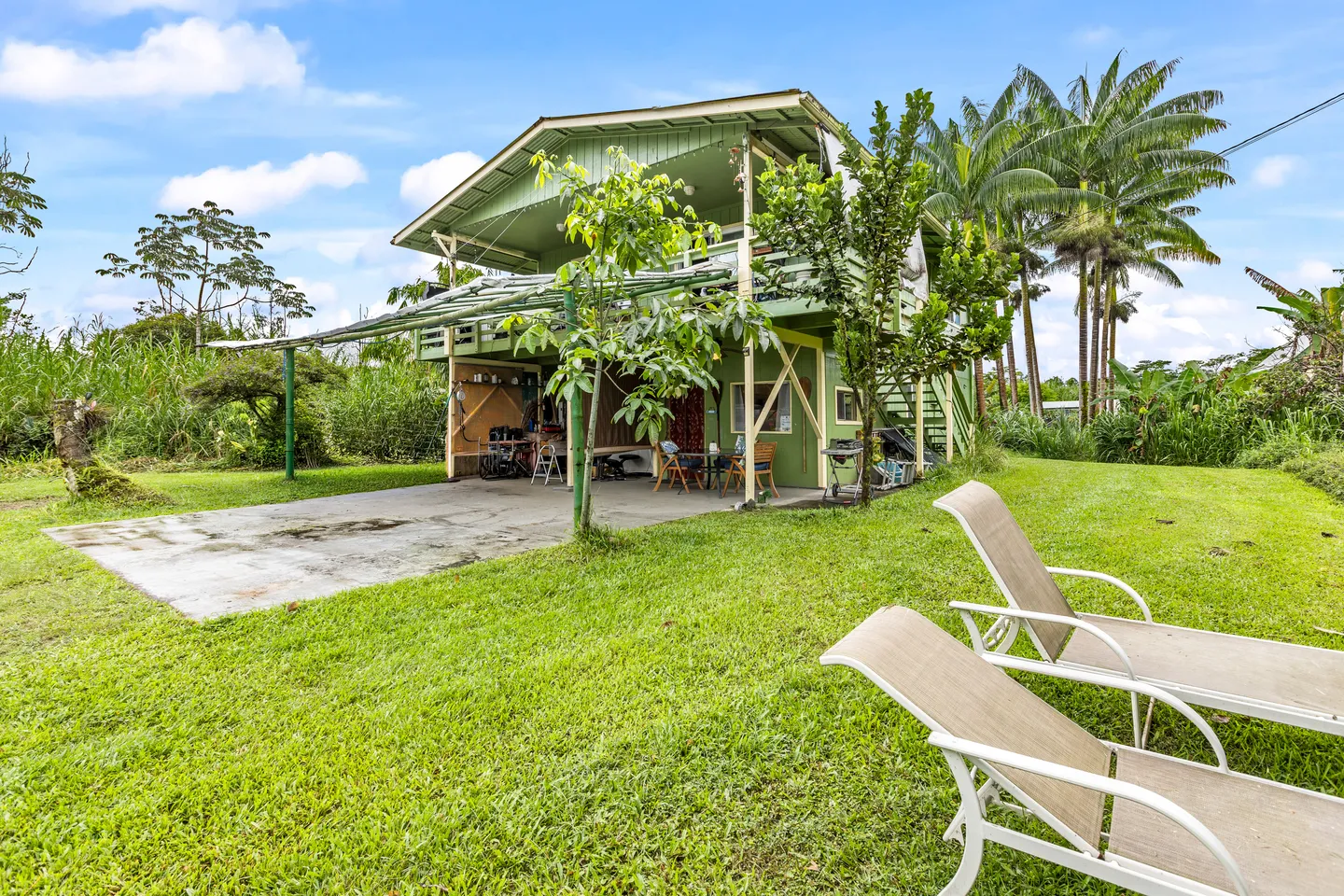 Exterior view of a two-story green house with a covered lower patio and balcony, surrounded by a lawn and palm trees with two lounge chairs in the foreground.