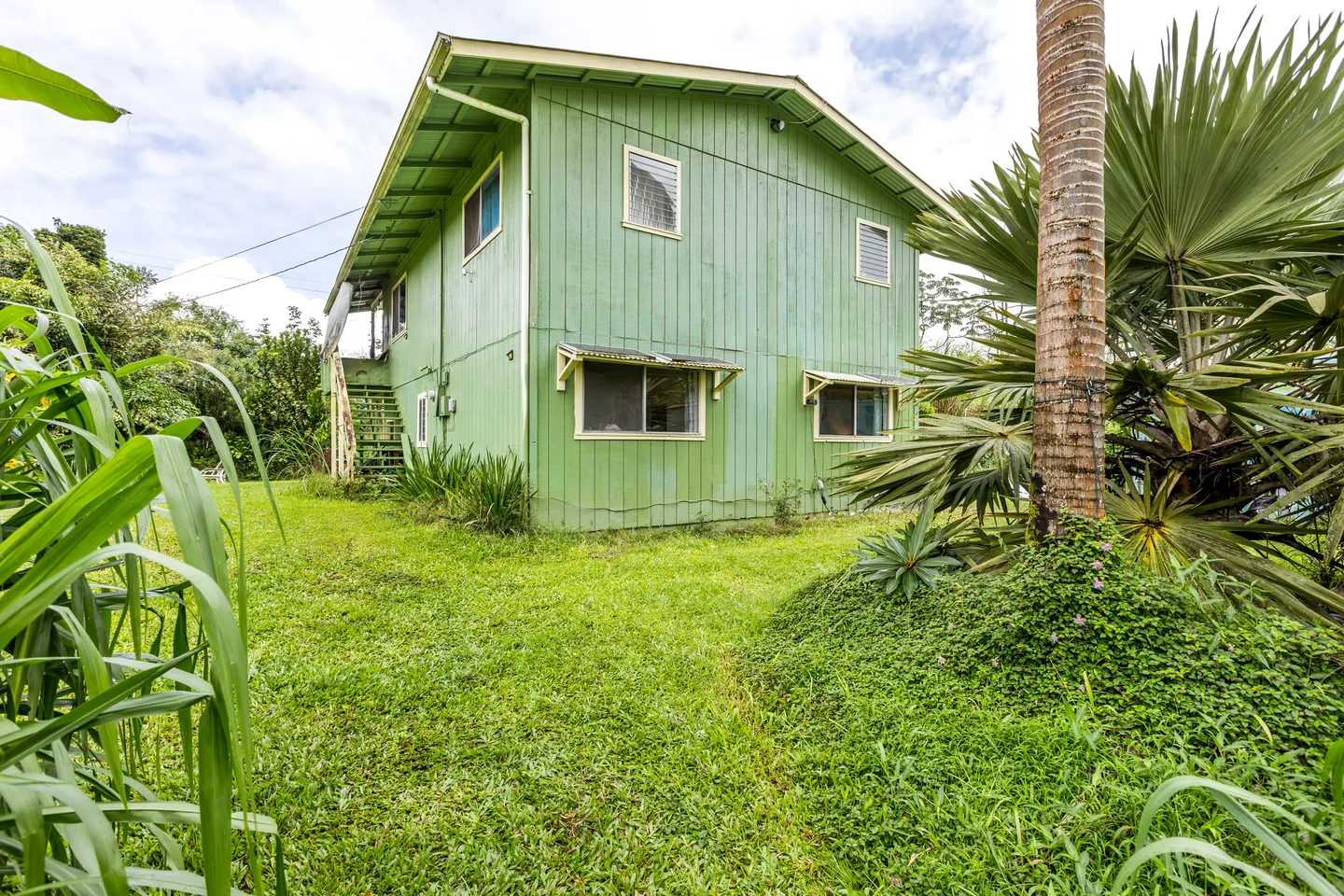 Exterior view of a two-story green-sided house surrounded by grass, palm trees, and tropical foliage.