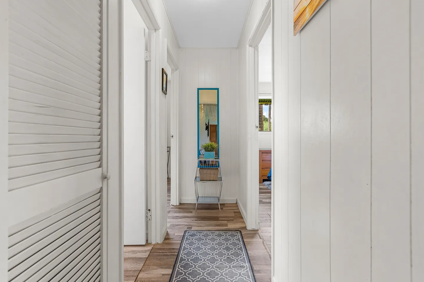 A brightly lit hallway featuring white paneled walls, wood-look flooring, a patterned runner rug, and a small decorative stand with a mirror.