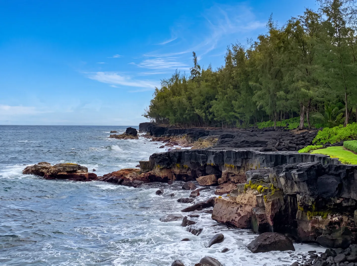 Rocky ocean coastline with dark volcanic rock shelves, crashing waves, and a dense line of green trees under a blue sky.