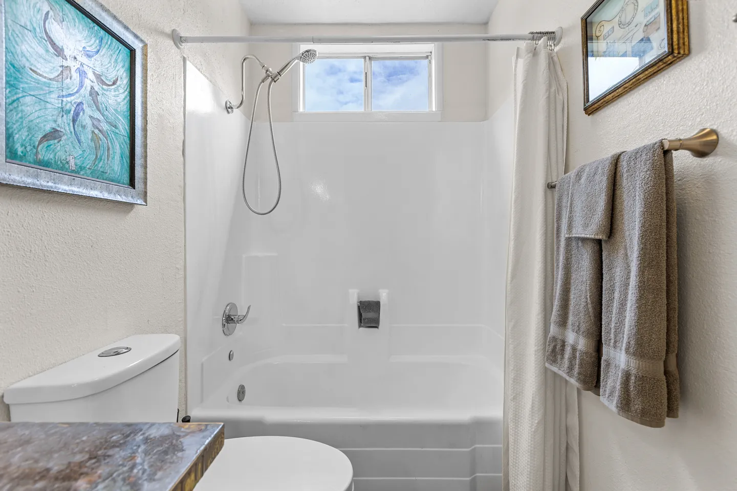 Bathroom with a white bathtub and shower combination, a small window above, a toilet, and a towel bar with towels.