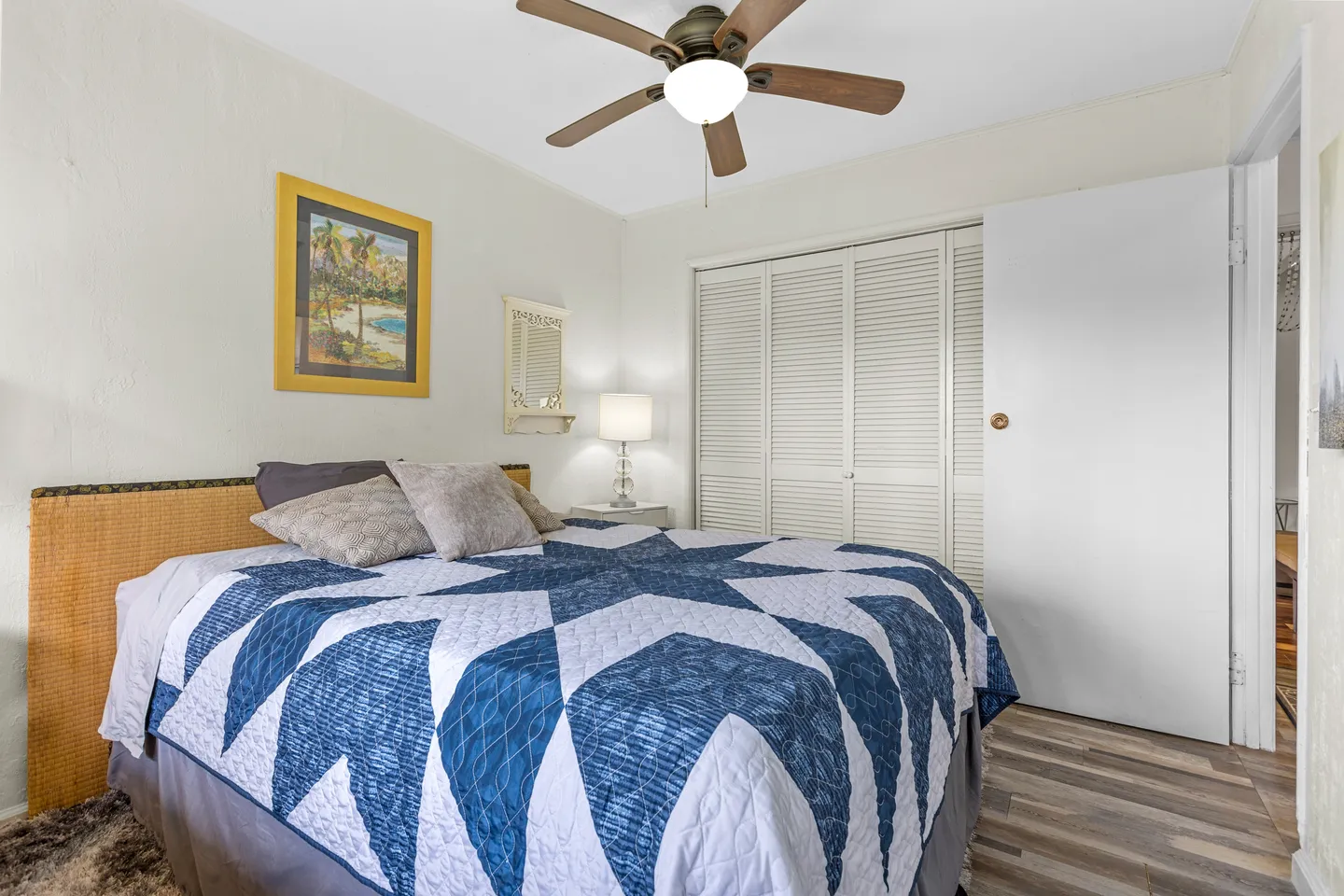 Bedroom with a bed made with a blue and white geometric quilt, wood-finish ceiling fan, and louvered closet doors.