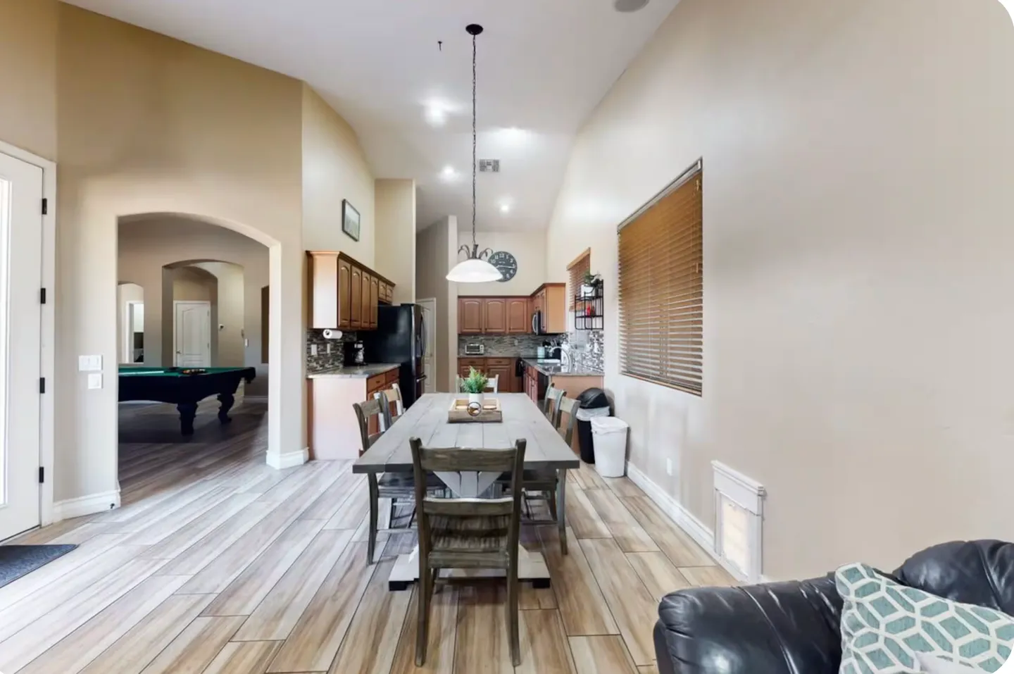 Dining area with a wooden table and chairs, leading into a kitchen with wood cabinetry and an arched doorway showing a pool table.