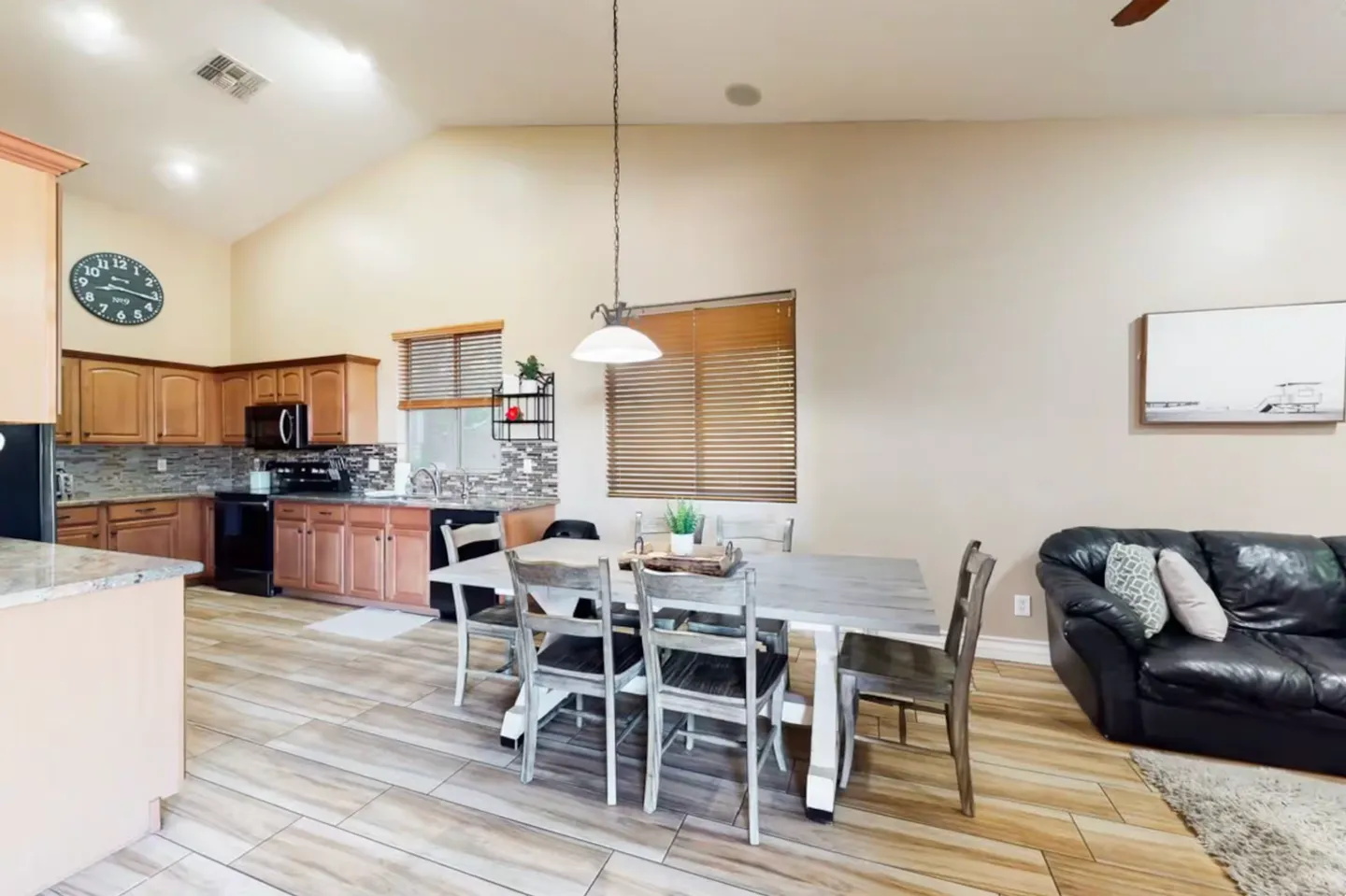 Open-concept dining area with a rustic dining table and chairs, adjacent to a kitchen with wooden cabinets and a living room with a black leather sofa.