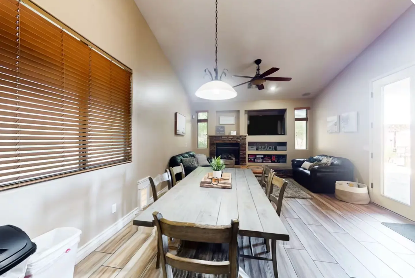 Dining area with a long wooden table and chairs leading into an open living room with a fireplace and television.