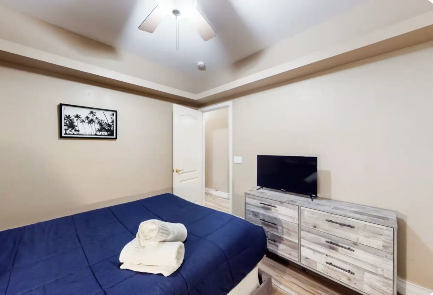 Bedroom featuring a bed with a blue comforter and folded white towels, a weathered wood dresser with a flat-screen TV, and an open doorway.