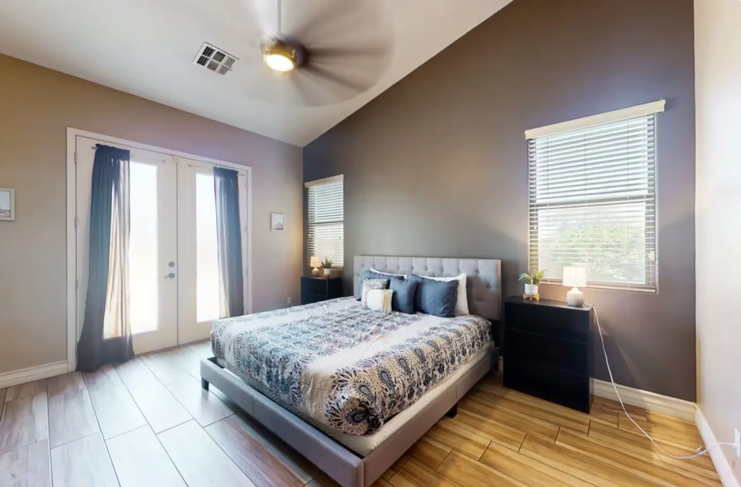 Bedroom with vaulted ceiling, wood-look tile flooring, gray tufted bed frame, and French doors.