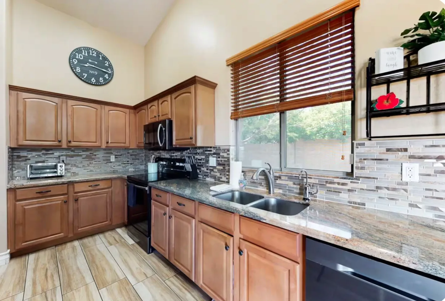 Kitchen corner featuring wooden cabinets, speckled countertops, a double basin sink beneath a window with blinds, and tile flooring.