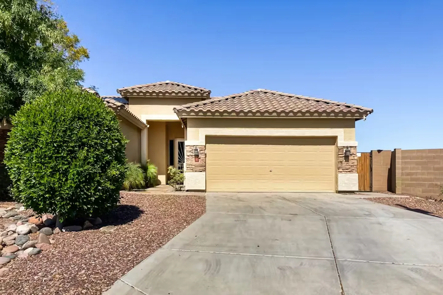 Exterior front view of a stucco single-story home featuring a clay tile roof, two-car garage, and concrete driveway under a clear blue sky.