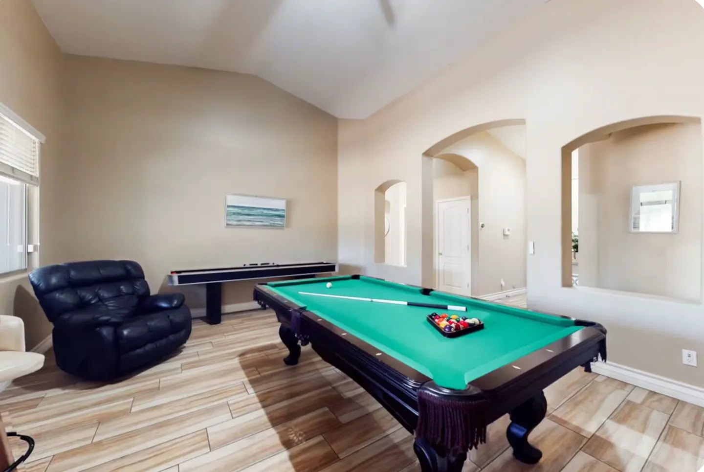 Game room featuring a green felt pool table, shuffleboard table, and black armchair on wood-look tile floors.