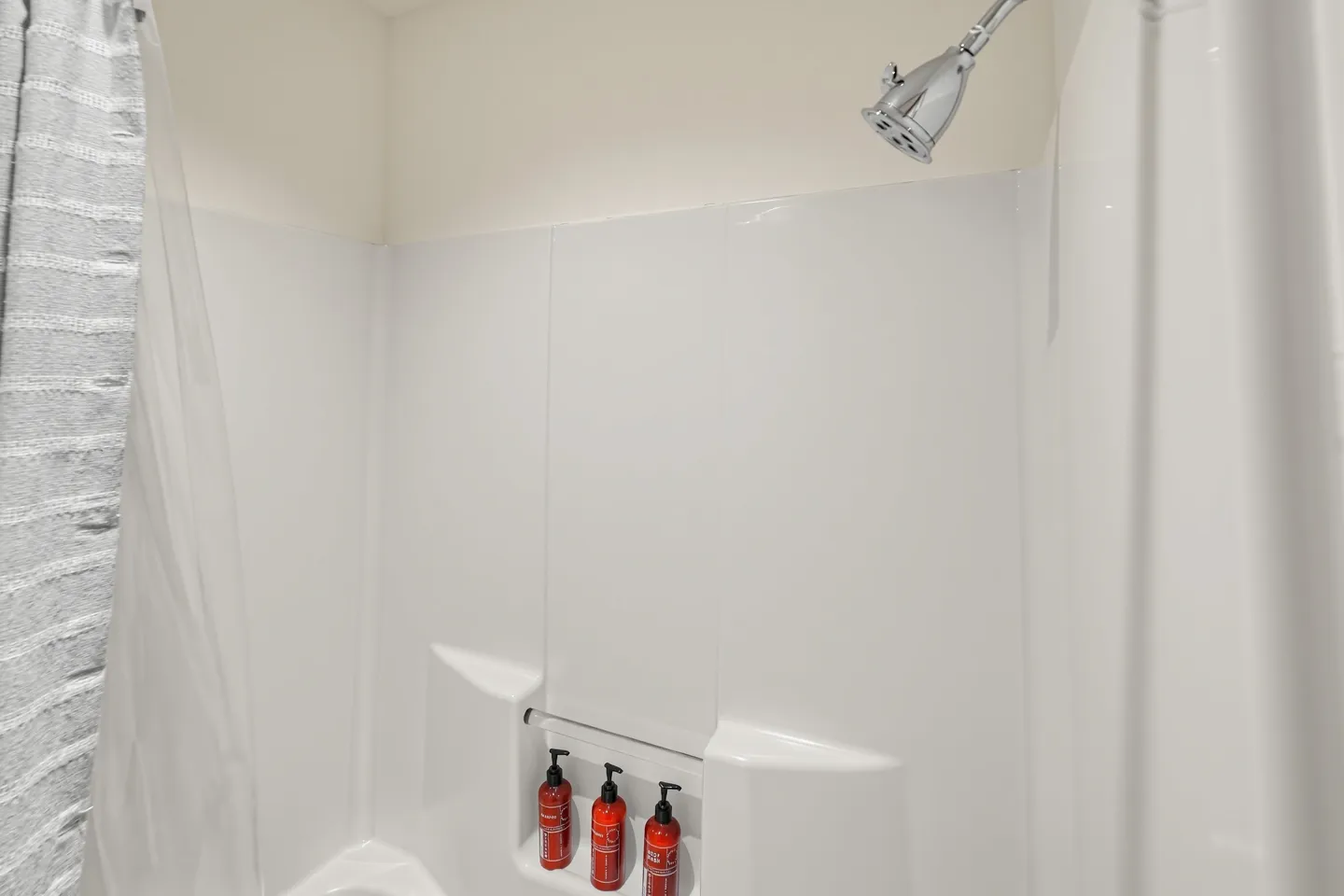 White tub and shower enclosure with chrome showerhead and red pump bottles on a built-in ledge.
