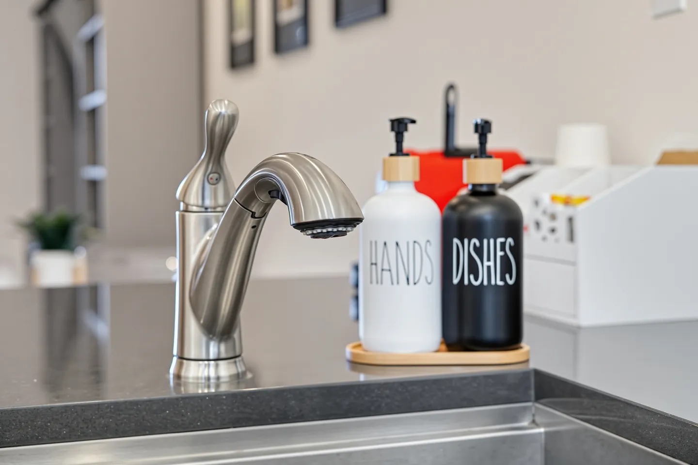 Close-up of a brushed metal kitchen faucet and dispenser bottles labeled HANDS and DISHES on a dark countertop.