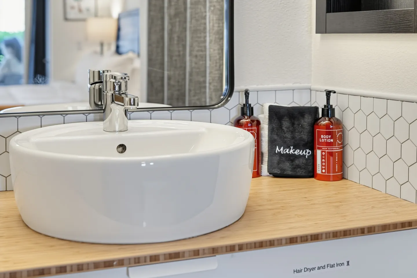 Close-up of a white vessel sink on a light wood vanity with a chrome faucet, black-framed mirror, and complimentary toiletries.
