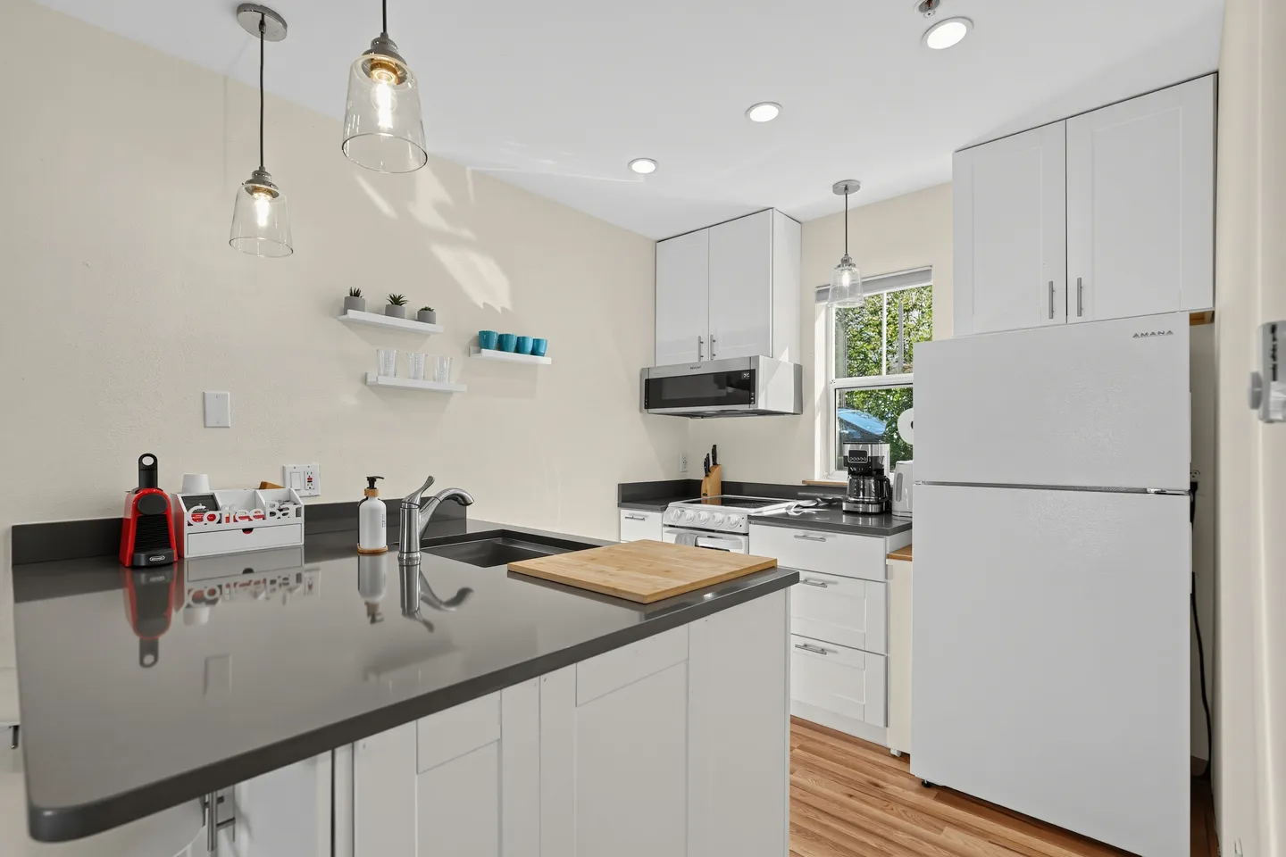 A compact kitchen featuring white cabinetry, dark grey countertops, pendant lights, and a white refrigerator.