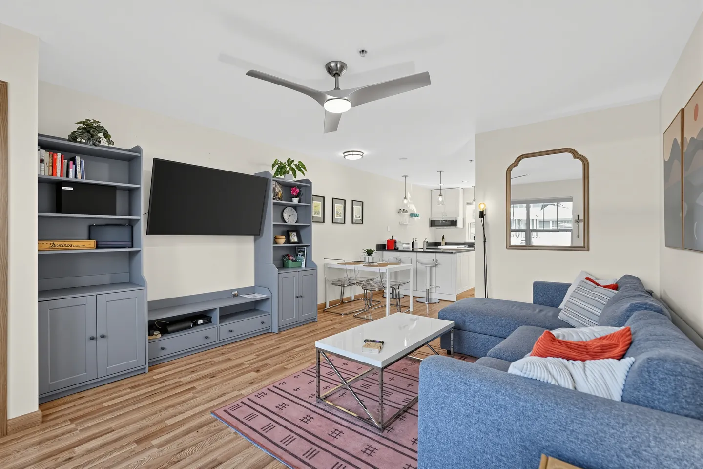 Open-concept living room with blue sectional sofa, wall-mounted television, blue entertainment center, coffee table, and view into the kitchen.