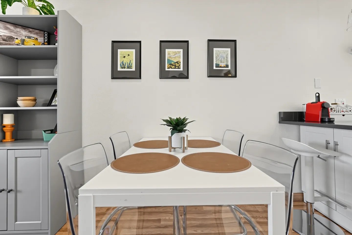 Dining area featuring a white rectangular table set with placemats and clear acrylic chairs, next to kitchen cabinetry.