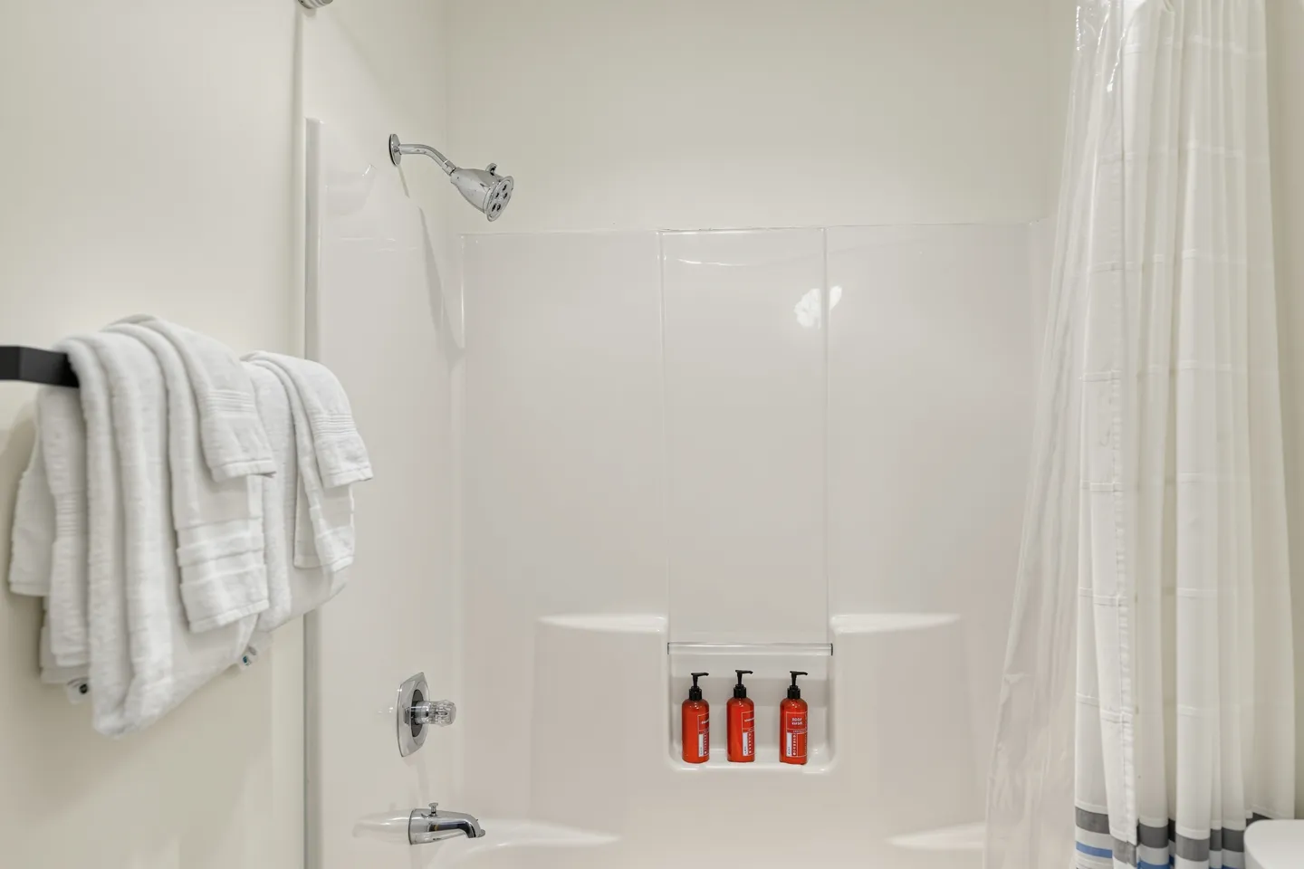 A clean white bathtub and shower combination with chrome fixtures, shampoo bottles on a built-in shelf, and folded white towels on a rack.