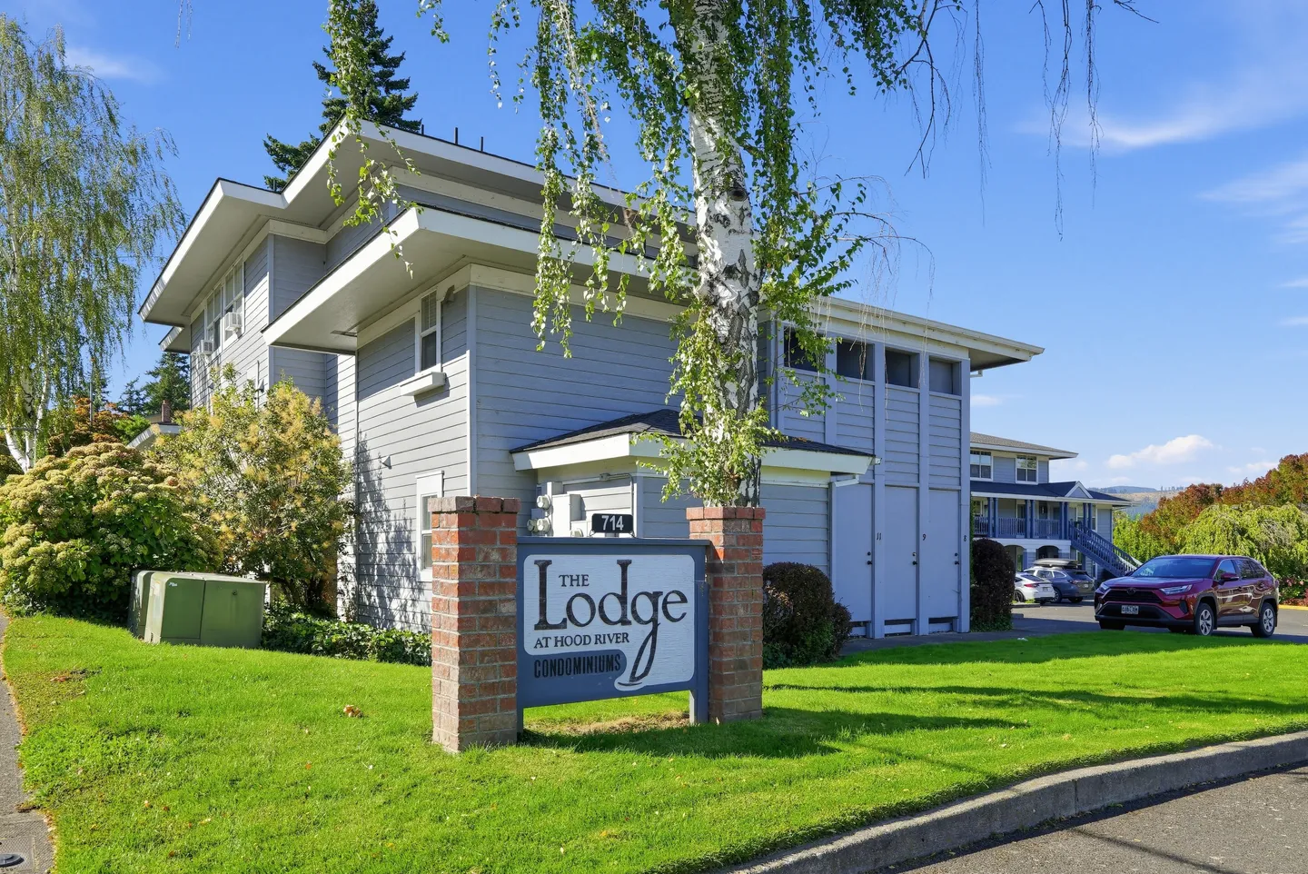 Exterior view of The Lodge at Hood River Condominiums sign set in brick pillars in front of multi-story buildings and parked cars.