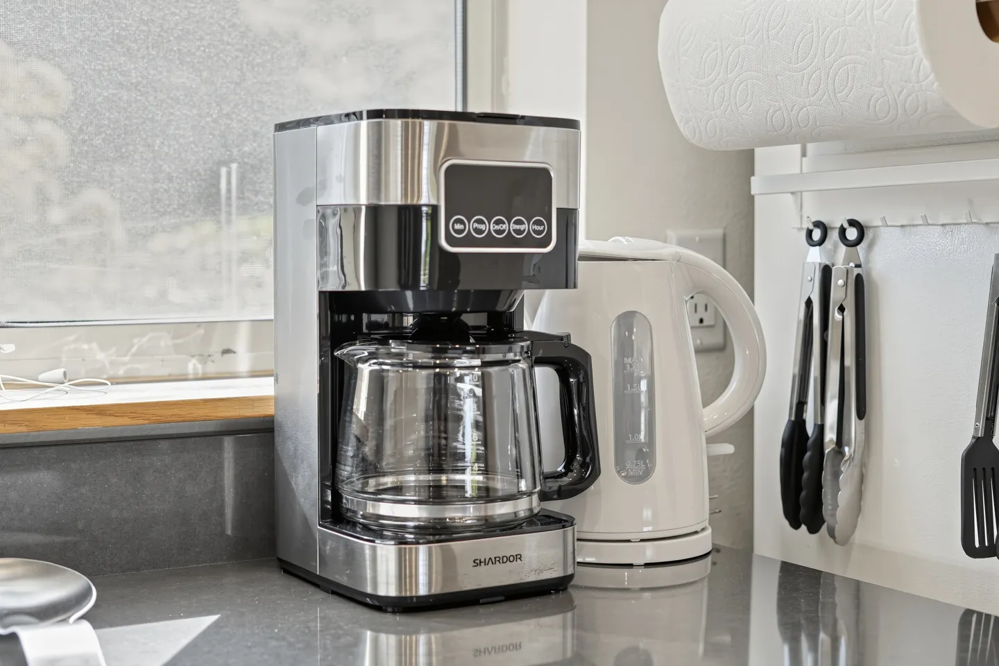 Close-up of a stainless steel drip coffee maker and a white electric kettle on a kitchen countertop.