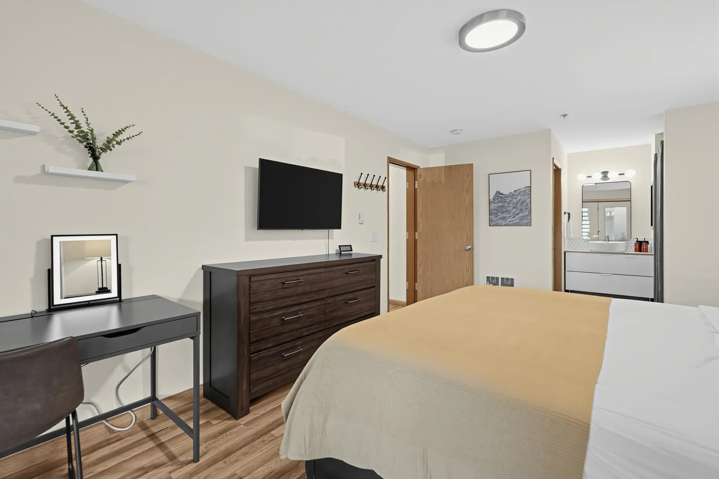 Bedroom featuring a bed with a tan blanket, a wooden dresser beneath a wall-mounted TV, a small desk with a vanity mirror, and an adjoining bathroom.