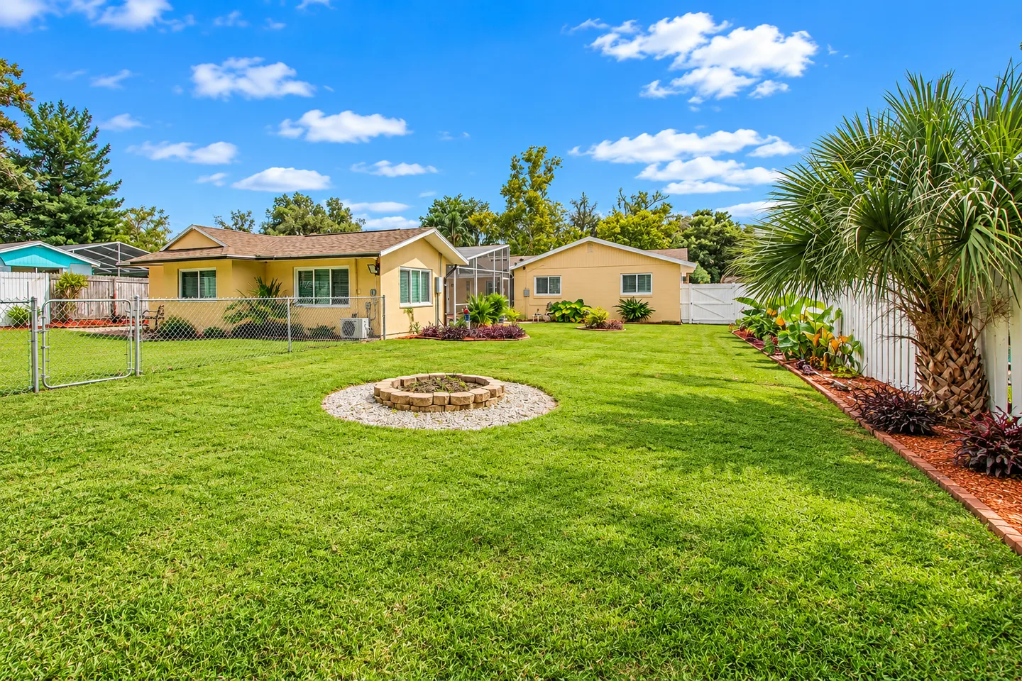 Spacious grassy backyard with a central stone fire pit, bordered by a white fence and palm trees, leading to yellow residential buildings under a bright blue sky.