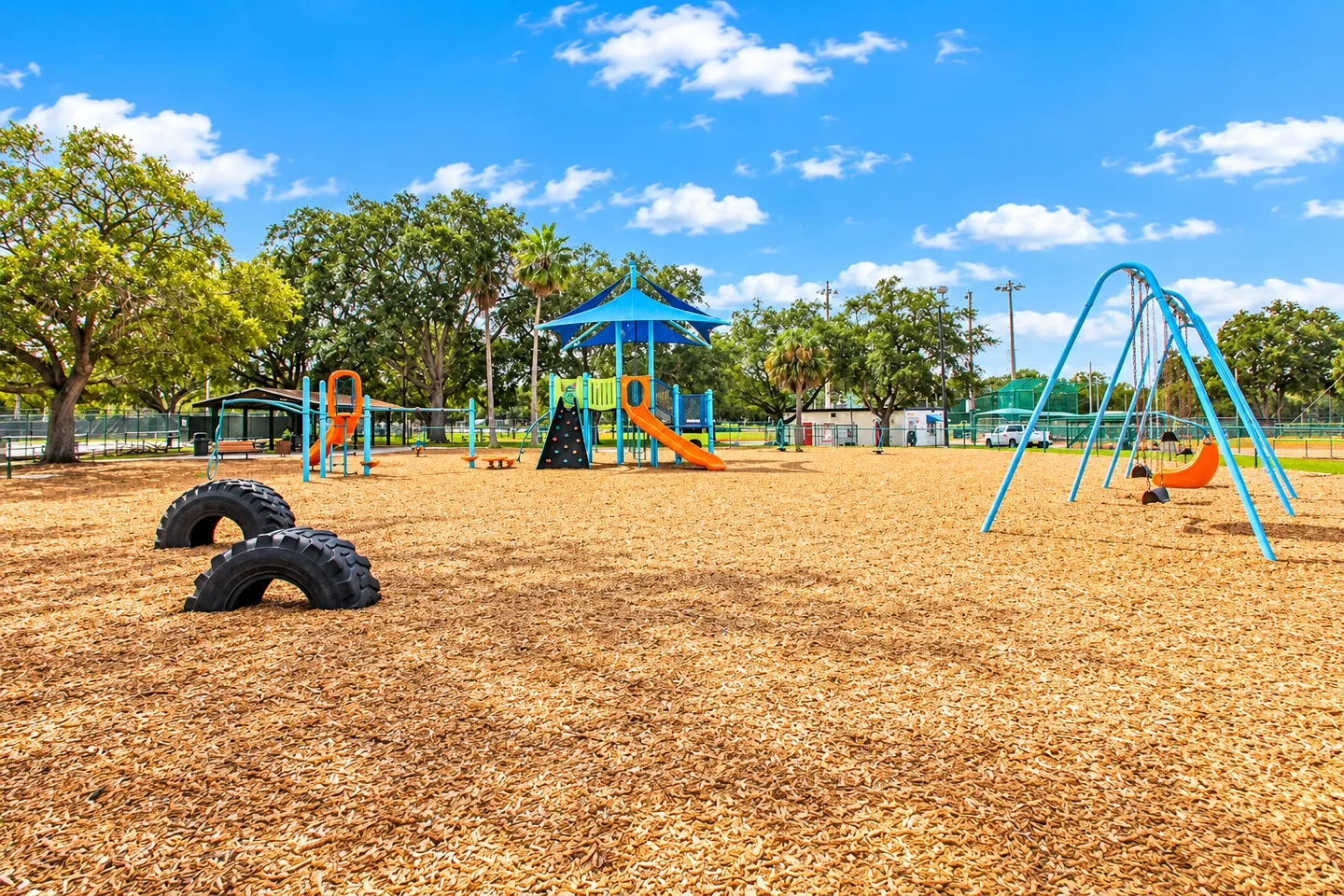 Community playground area with wood chip ground cover, swing set, slides with shade canopy, and embedded tires under a bright blue sky.
