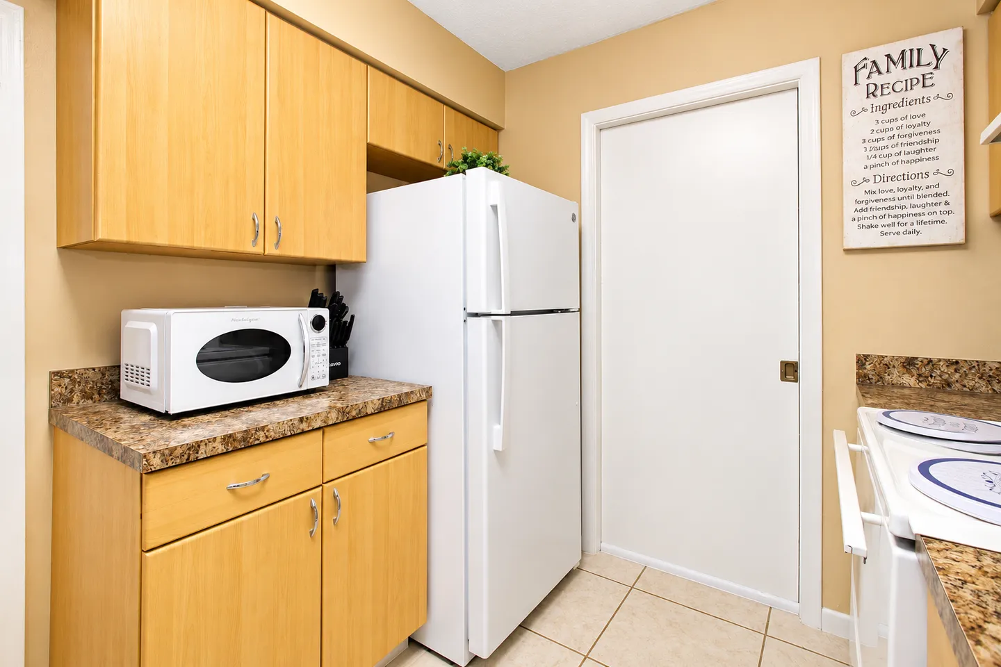 Kitchen area showing light wood cabinets, countertop with microwave, white refrigerator, and tiled floor.