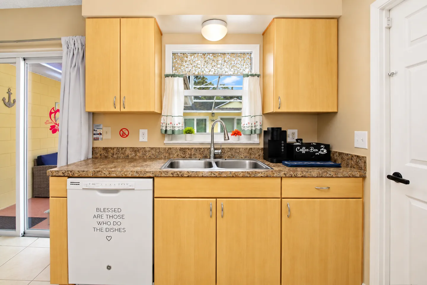 Kitchen counter area with a double sink, light wood cabinets, white dishwasher, and window looking outside.