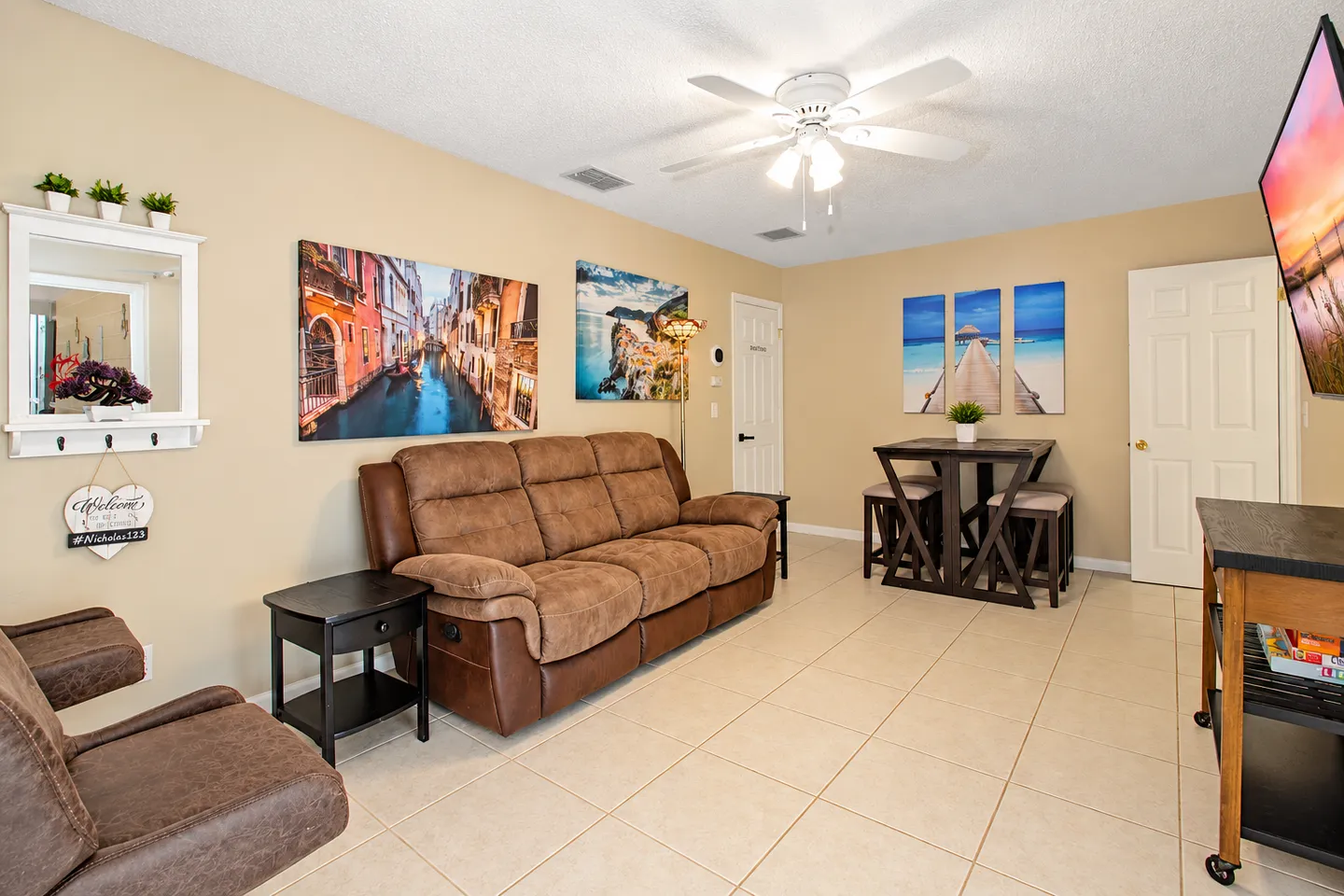Living room with brown reclining sofa, tile floors, a small high-top dining table with stools, and wall art.