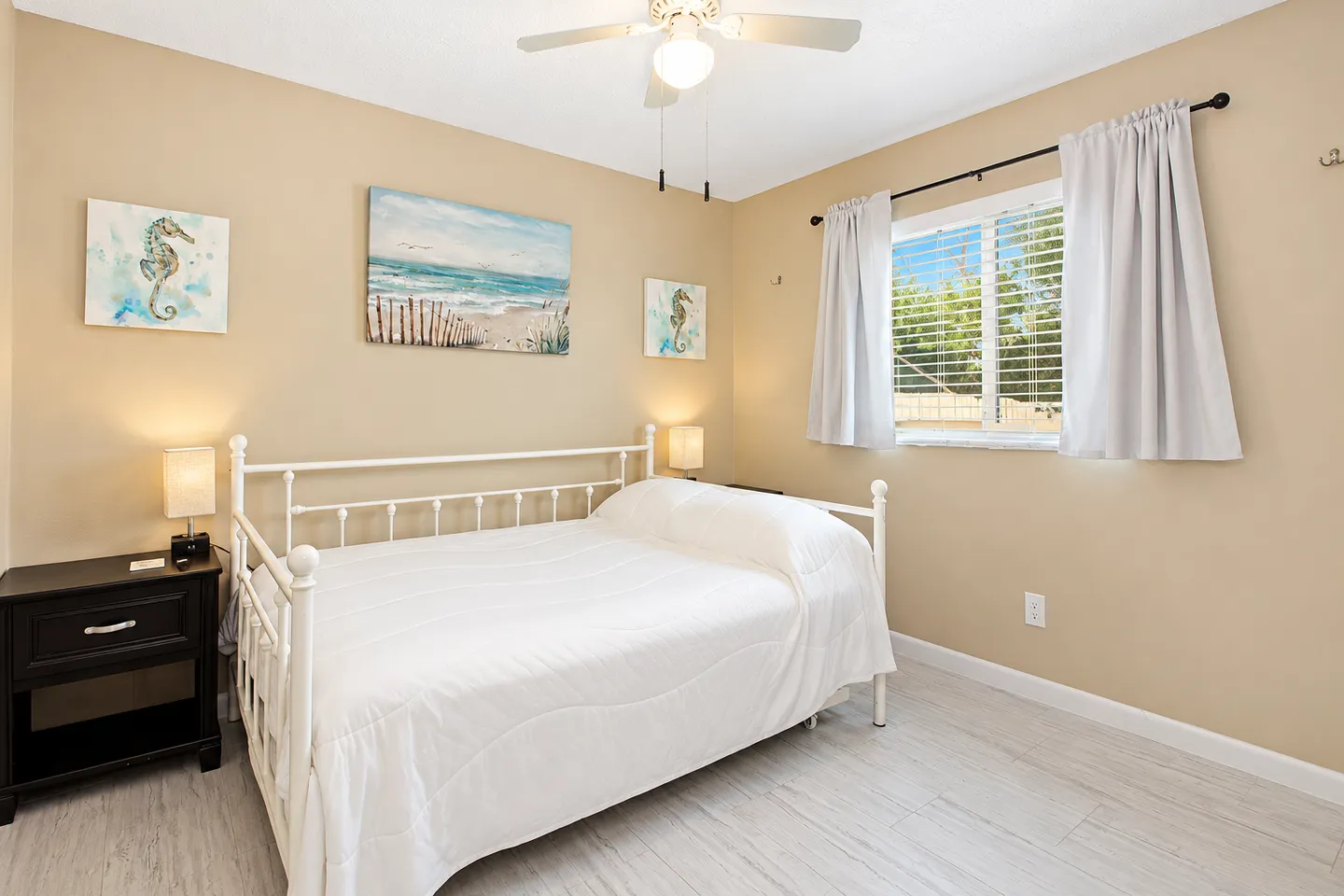 Bedroom with a white metal daybed, light wood-look flooring, beige walls, and coastal-themed wall art.