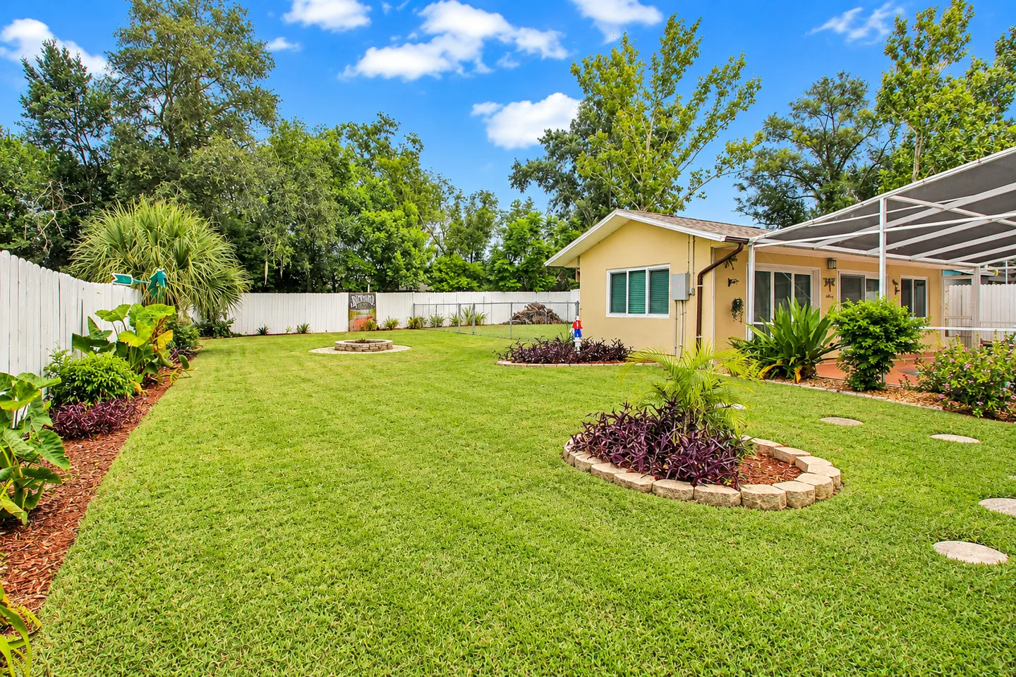 Spacious grassy backyard with landscaped garden beds, a fire pit, and white fencing alongside a single-story house.