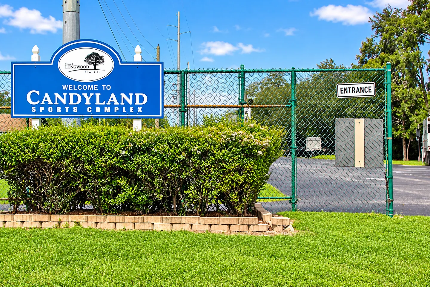 Blue sign reading Welcome to Candyland Sports Complex in front of a green chain-link fence and lawn.