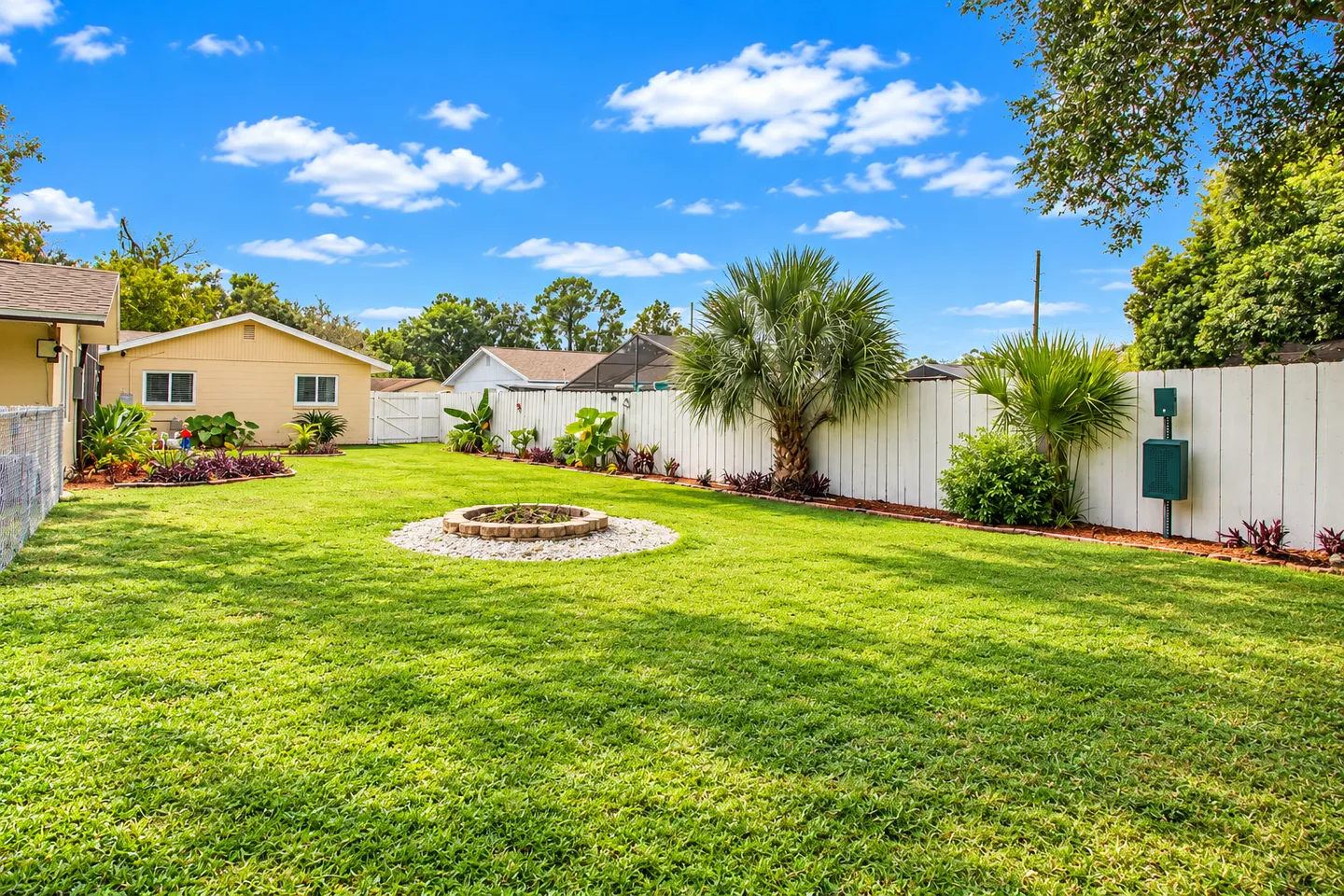 Spacious grassy backyard enclosed by a white fence, featuring a stone fire pit in the lawn and palm trees along the perimeter under a bright blue sky.