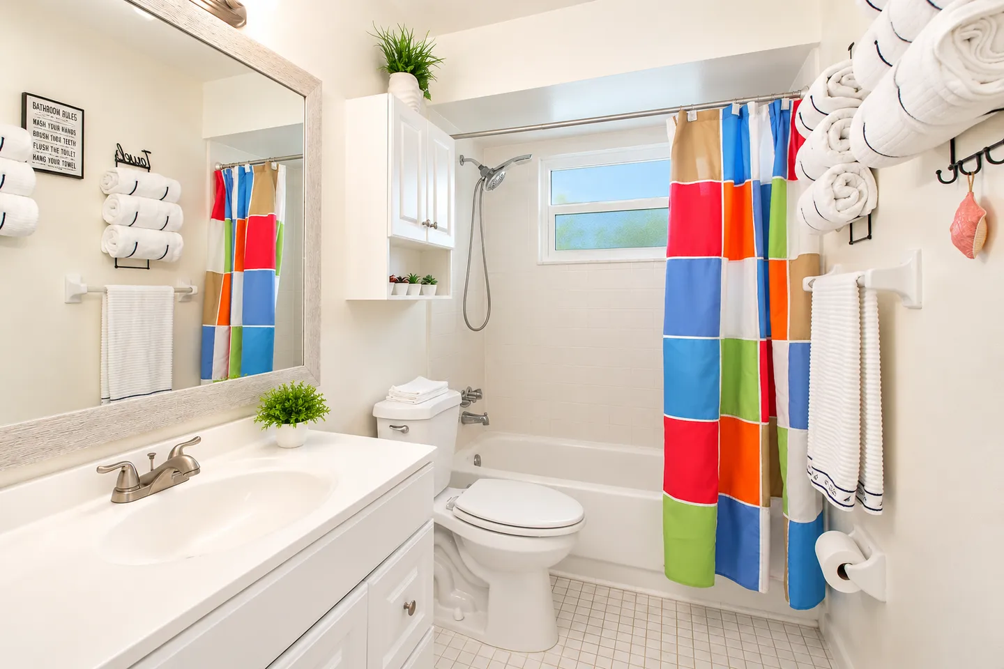 Bright bathroom featuring a white vanity, framed mirror, toilet, shower and tub combination with a colorful checkered curtain, and towel racks.