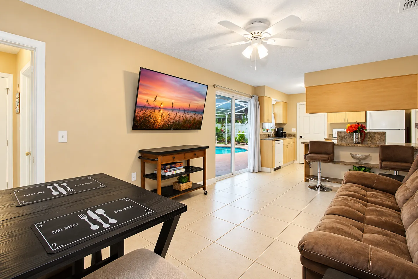 Open-concept living and dining area with tile flooring, a wall-mounted TV, brown sofa, and sliding glass doors leading to a pool patio.
