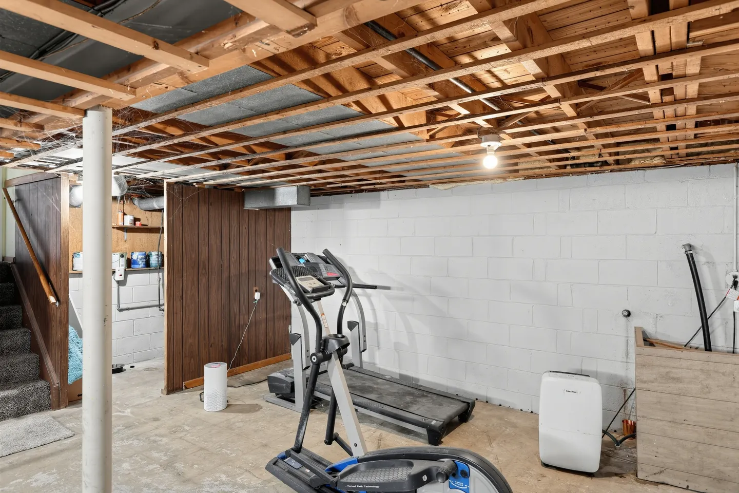 Unfinished basement area with painted cinder block walls, exposed ceiling framing, a treadmill, an elliptical machine, and a dehumidifier on a concrete floor.