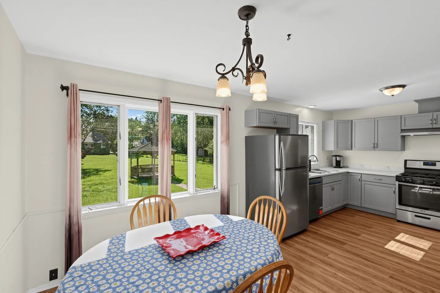 Dining area with a round table and chairs in the foreground, next to a kitchen with grey cabinetry and stainless steel appliances, with windows overlooking a grassy yard.