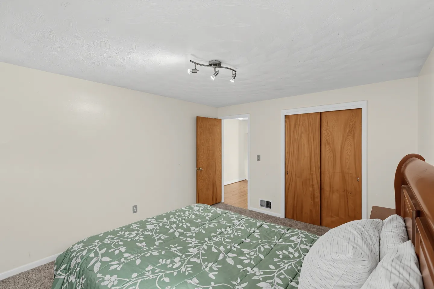 Bedroom with carpet, neutral walls, a bed with green leaf-patterned bedding, track lighting, and wood doors.
