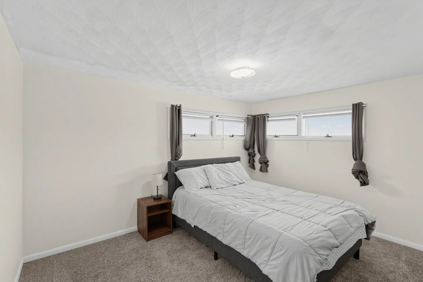 Bedroom with a bed made with gray bedding, a wooden nightstand, carpeted flooring, and high horizontal windows with tied curtains.