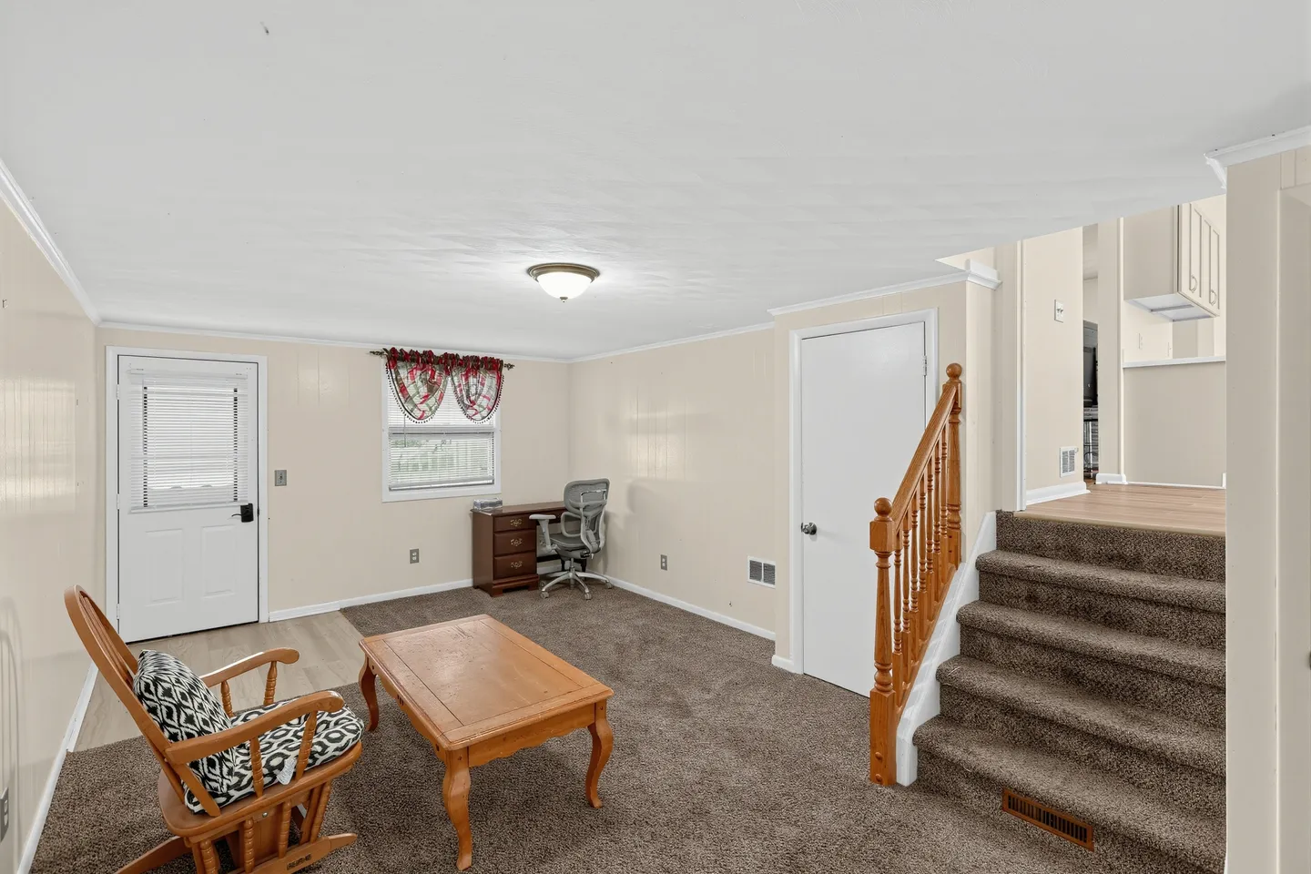 Lower-level living area with brown carpeting, a wooden coffee table, rocking chair, desk with office chair, and carpeted stairs leading up.