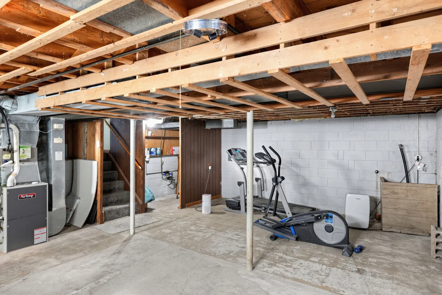 Unfinished basement with concrete floor, exposed wooden ceiling joists, exercise equipment, furnace, and stairs.