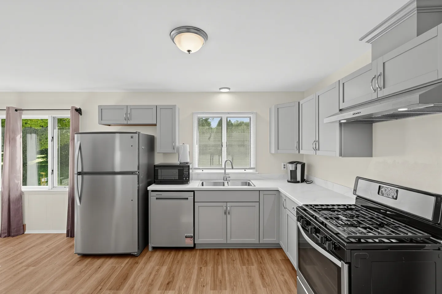 Bright kitchen featuring light gray cabinetry, stainless steel appliances, white countertops, and wood-look flooring.