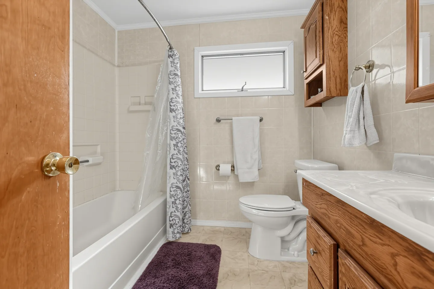 Bathroom with a bathtub shower combination, tiled walls, toilet, vanity with wooden cabinetry, and small window.