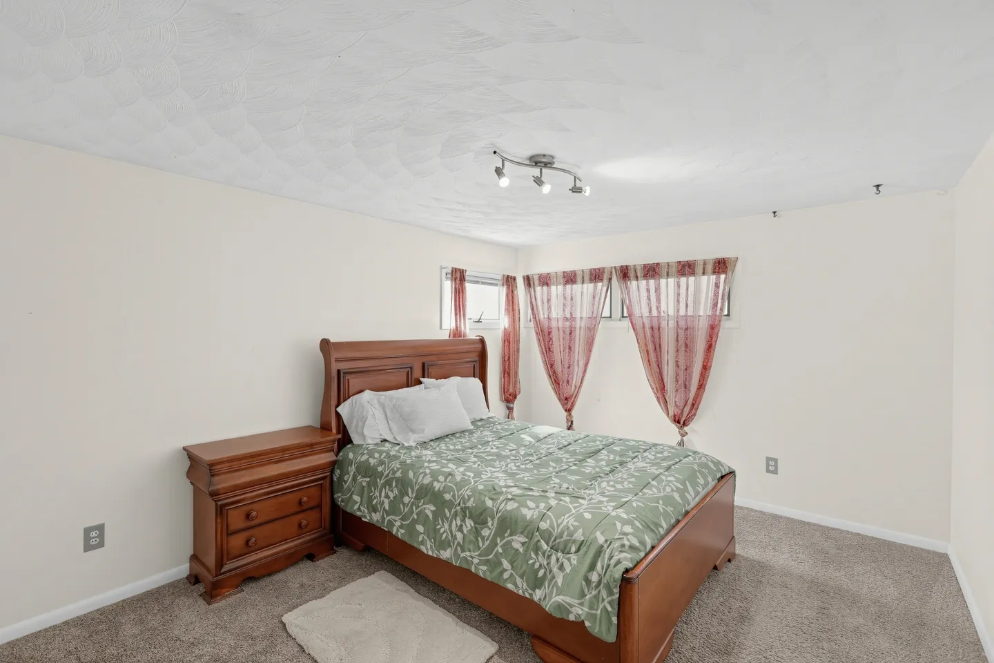 Bedroom with a wood frame bed, matching nightstand, textured ceiling, and windows with red-toned curtains.