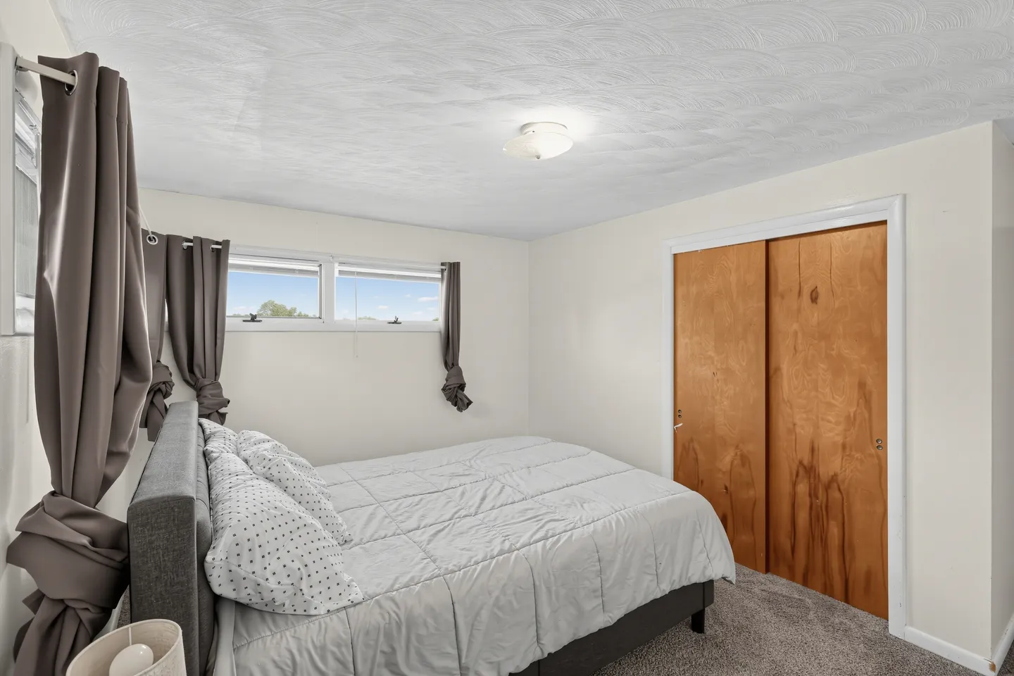 Bedroom with a made bed, carpeted flooring, small windows with gray curtains, and wooden sliding closet doors.