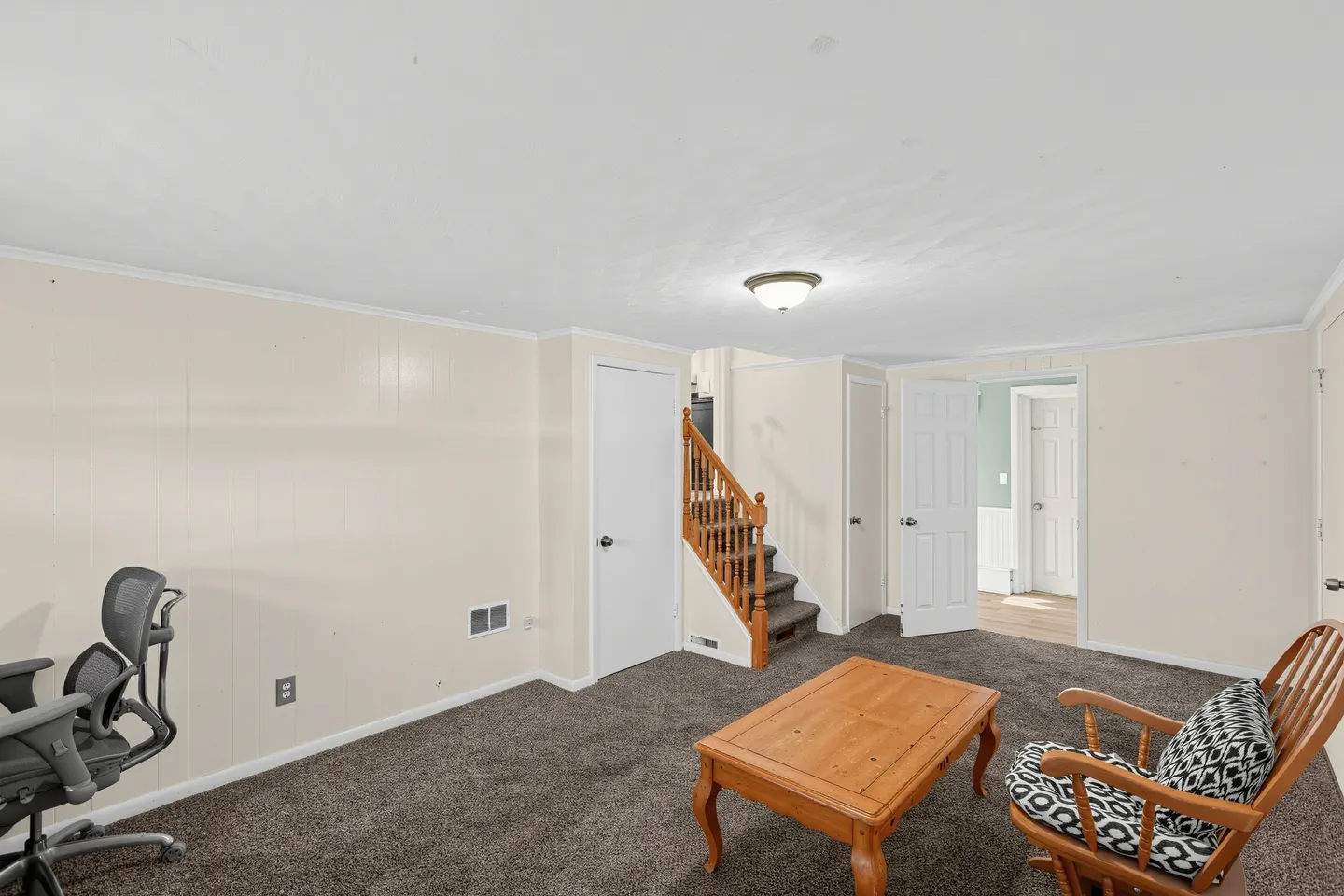 A carpeted indoor living space with paneled walls, a wooden coffee table, a rocking chair, an office chair, and carpeted stairs leading up.