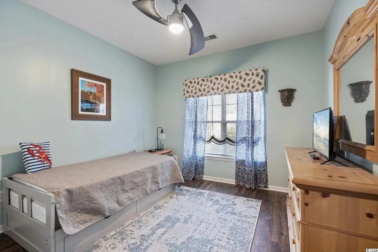 Bedroom featuring a daybed with trundle, light blue walls, wooden dresser with a TV and mirror, area rug, and window with patterned curtains.