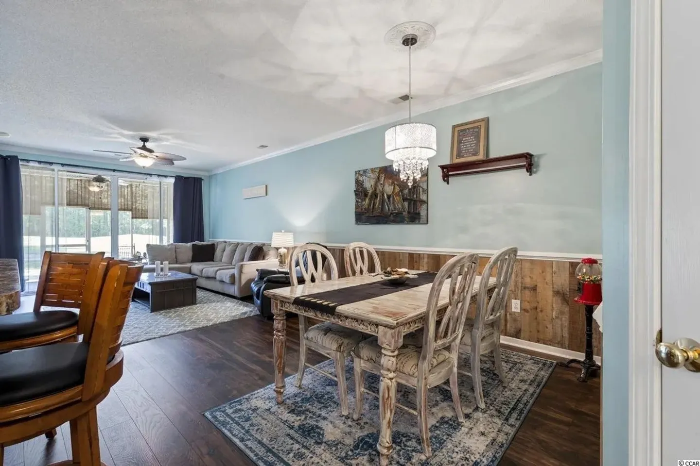 Dining area with a distressed wooden table and chairs opening to a living room with a sectional sofa and sliding glass doors.
